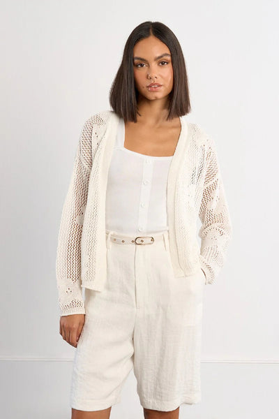 White fine knit open-front cardigan with floral openwork detail, long sleeves, and relaxed cropped fit, styled with cream linen shorts and a white tank — front view.