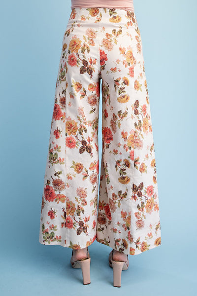 Model is wearing floral print wide leg pants in ivory with pink and yellow rose print, wide leg silhouette and seam details, back view.