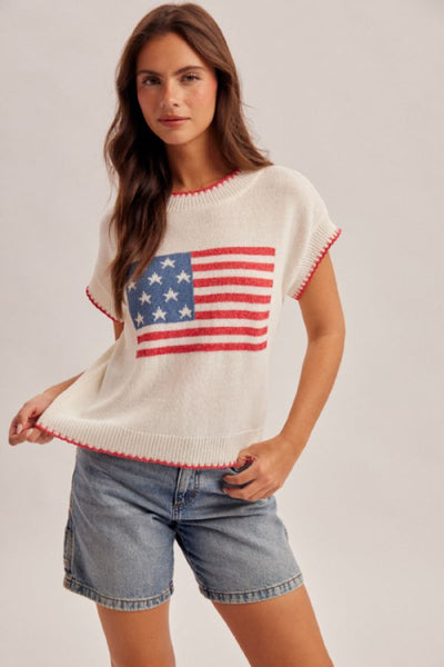 Cream lightweight knit sweater top with an American flag intarsia graphic on the front and red contrast stitching at neckline and sleeves, styled with denim shorts.