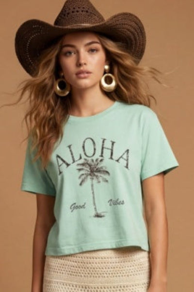 Model is wearing Aloha Wide Boy Tee in mint green with a palm tree graphic, styled with a crochet midi skirt and cowboy hat.